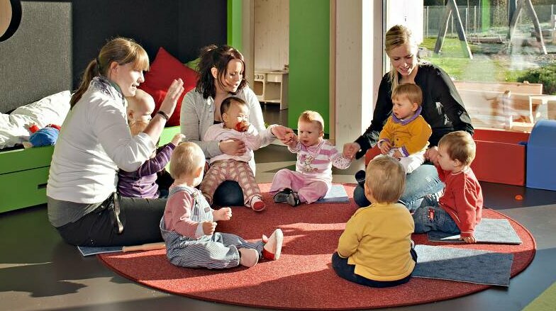 Blick in den Aufenthaltsraum einer Kita mit Spielgeräten und einer großen Fensterfront. Fünf Kinder sitzen mit zwei Erzieherinnen in einem Kreis