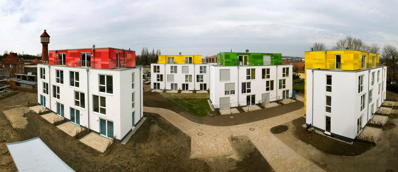Studentenwohnheim Osnabrück: vier weiße, quadrtische Häuser mit großen Fenstern und drei Etagen. In den höchsten Stockwerken gibt es Balkone, deren Fassaden in jedem Gebäude unterschiedlicher Farben sind: rot, gelb oder grün.