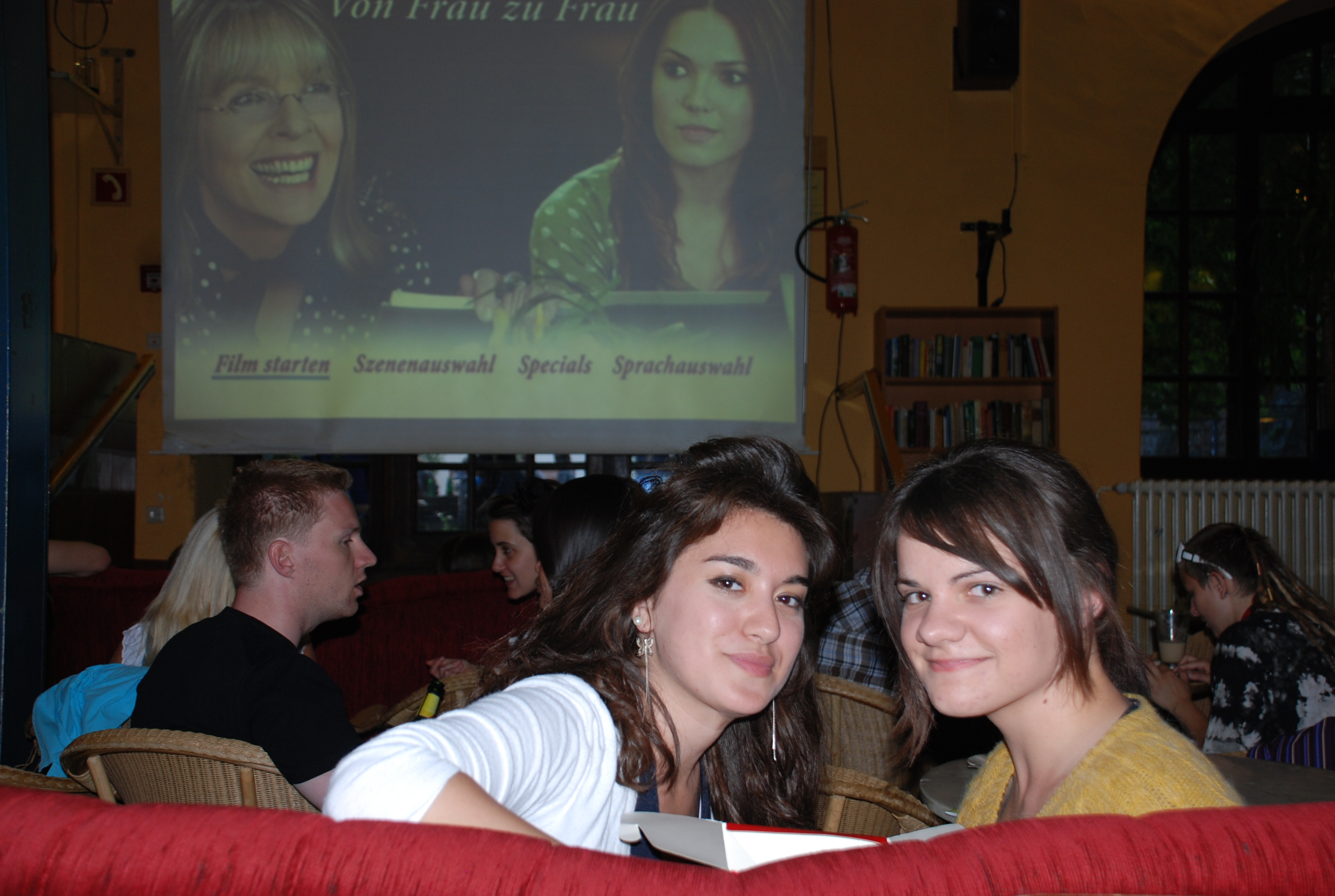 Zwei Mädels sitzen in einem Studentenclub auf einem Sofa. Hinter ihnen hängt eine große Leinwand auf der ein Film "Von Frau zu Frau" gezeigt wird.