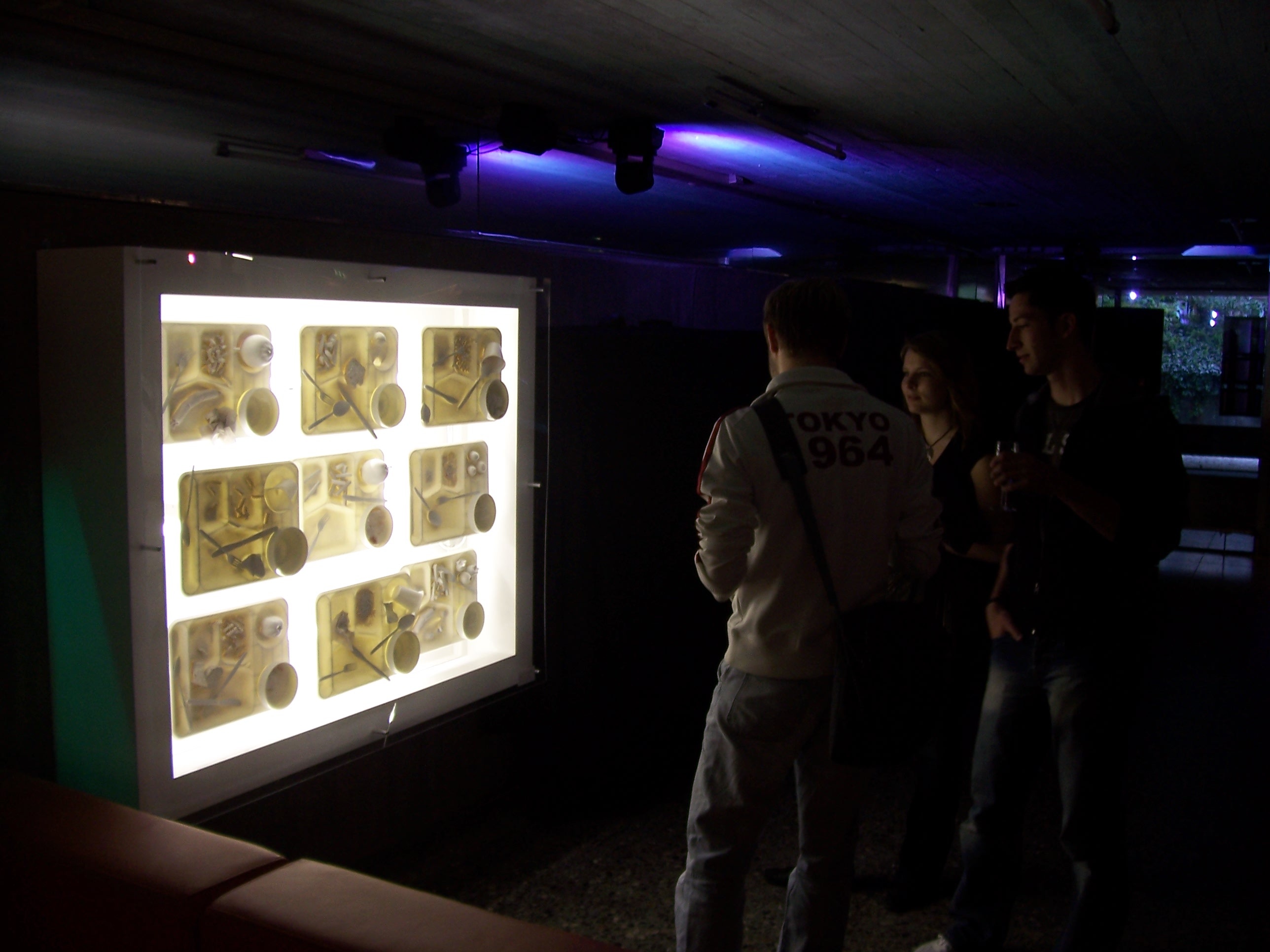 Drei Studenten betrachten ein Leuchtbox in einer Freiburger Mensa.  Darin: Kunstwerke aus Esstabletts.