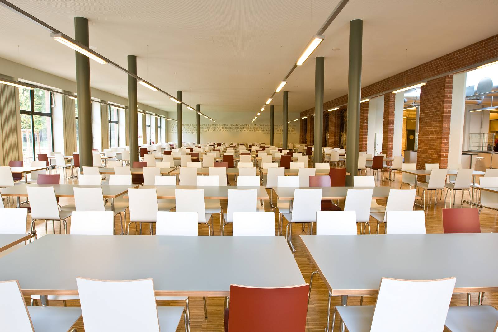 Der leere Essenssaal einer Mensa: Viele Tischreihen stehen in einem langen Saal. Von einer großen Fensterfront fällt viel Licht herein