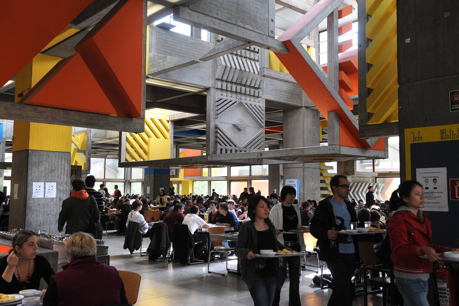 Blick in den Essenssaal einer Mensa, die Tische sind voll belegt, es laufen Studierende mit Essenstabletts durch die Gänge. Die Architektur ist aus Beton, dennoch bunt und modern.