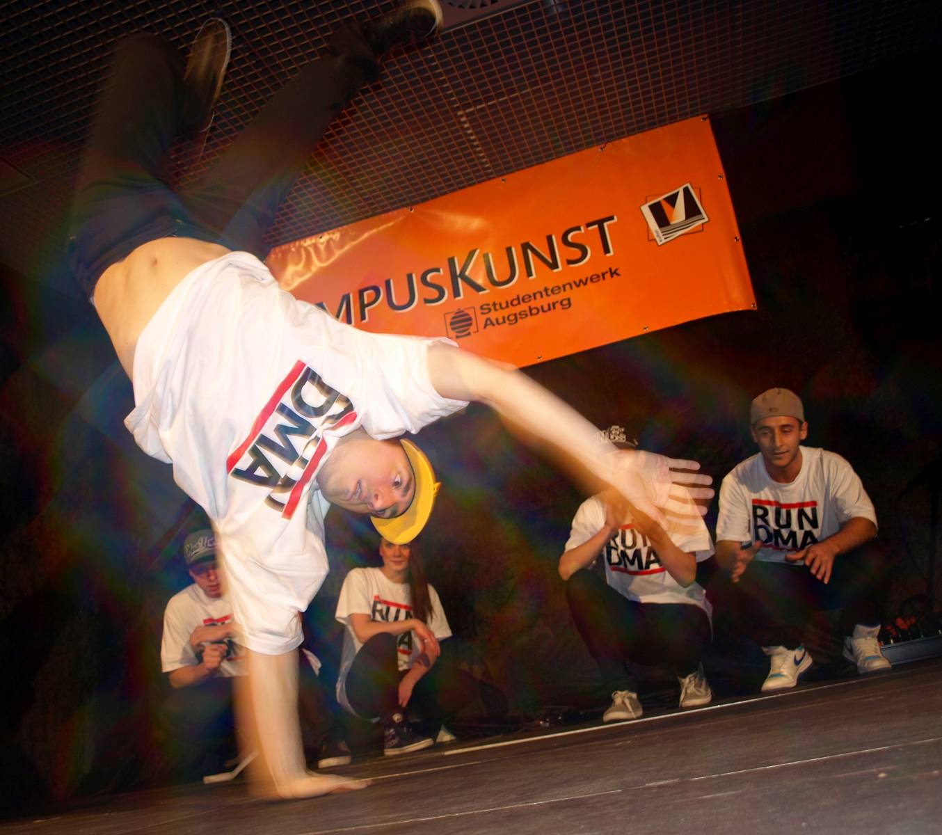 Eine Breakdancer-Gruppe beim Auftritt, alle in "RUN DMA" T-Shirts. Einer der Tänzer steht gerade nur auf einer Hand, die anderen schauen konzentriert zu.