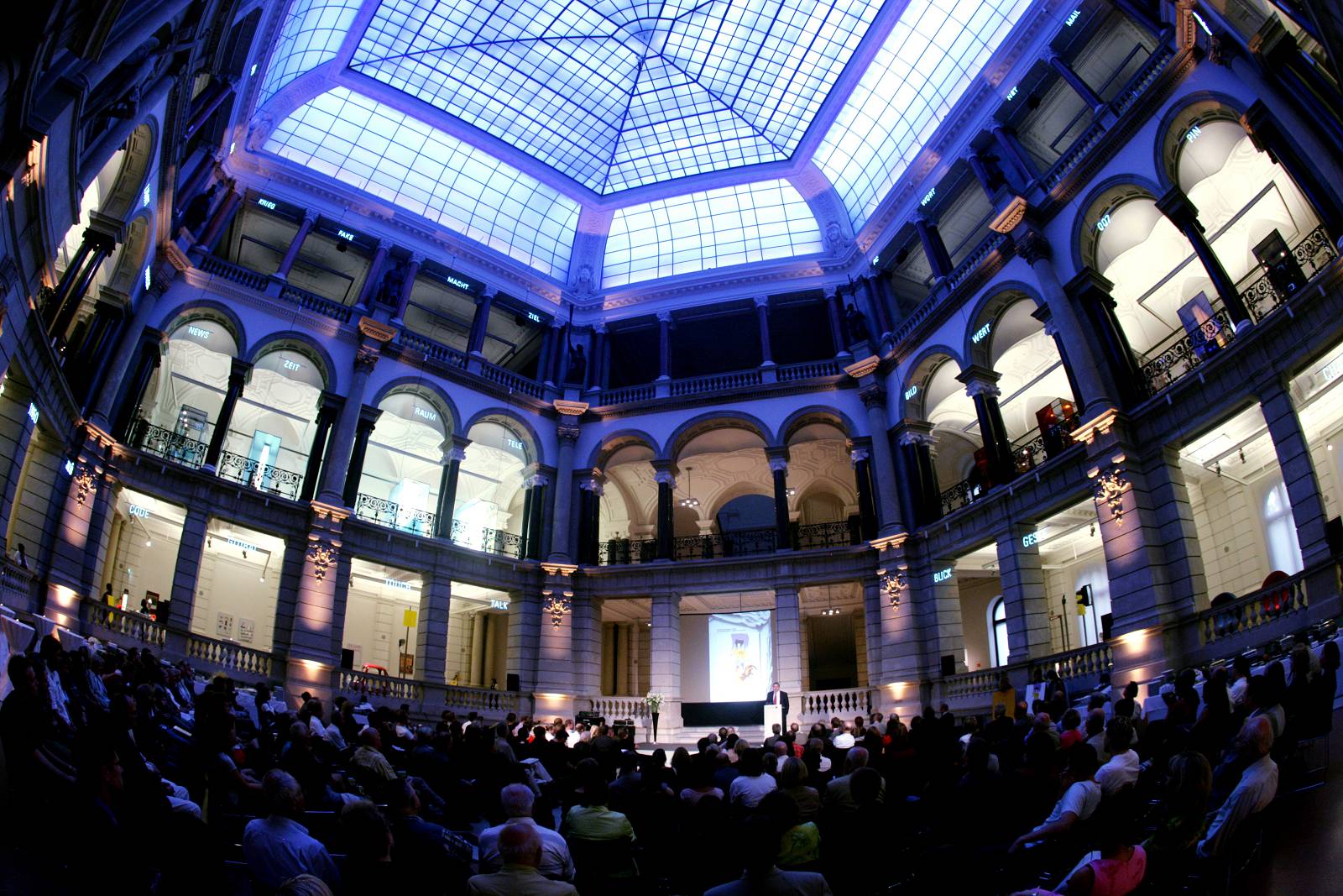 Panoramaaufnahme des Innenhofs im Museum für Kommunikation. Circa 200 Menschen sitzen im Publikum und gucken zur Bühne