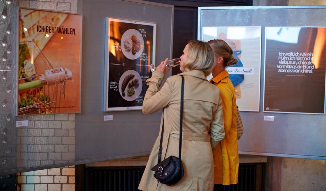 Zwei Frauen stehen vor einem Plakat der Ausstellung und trinken Sekt.