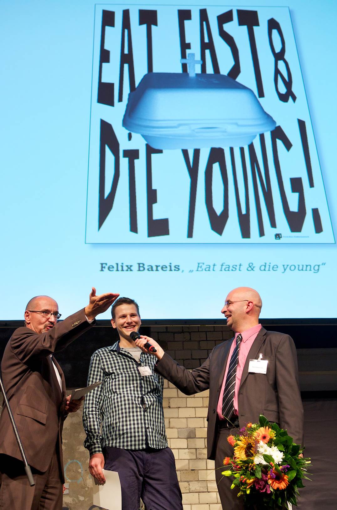 Felix Bareis (Mitte) mit einem seiner beiden Siegerplakate, umrahmt  von den Moderatoren | (c) Kay Herschelmann