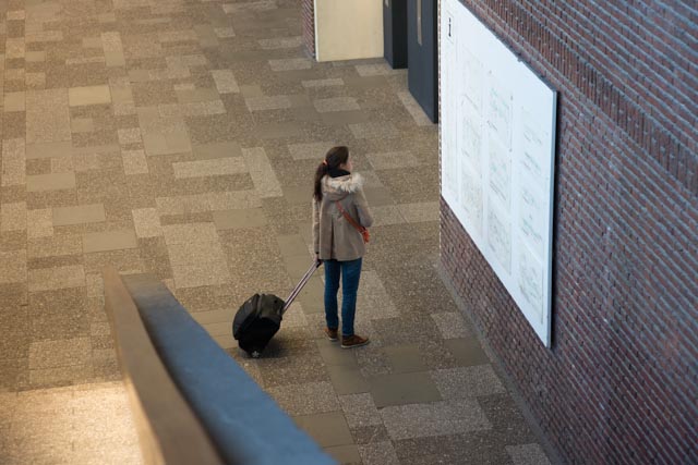 Studentin mit Rollkoffer steht in der leeren Eingangshalle einer Universität und betrachtet den Raumplan