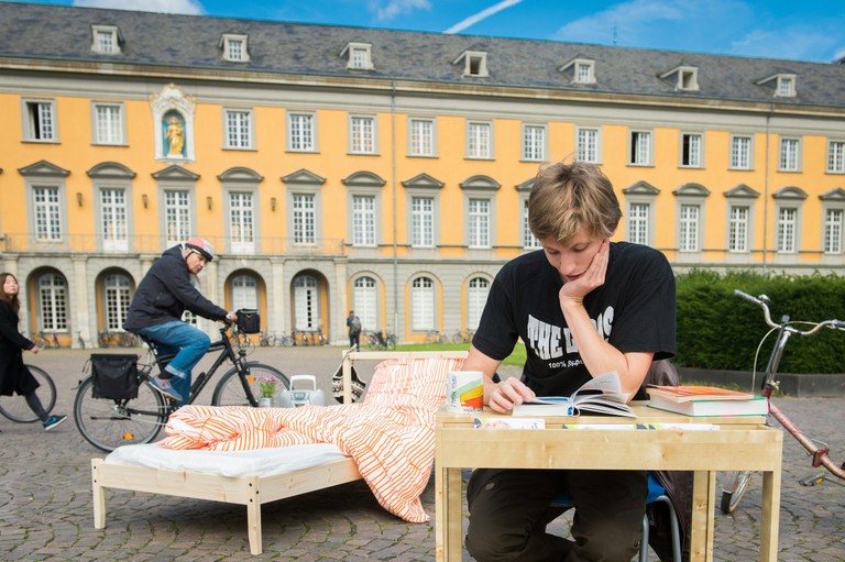 Tisch und Bett auf dem Bonner Hofgarten. Am Schreibtisch sitzt ein Student und liest. Im Hintergund fahren Radfahrer vorbei