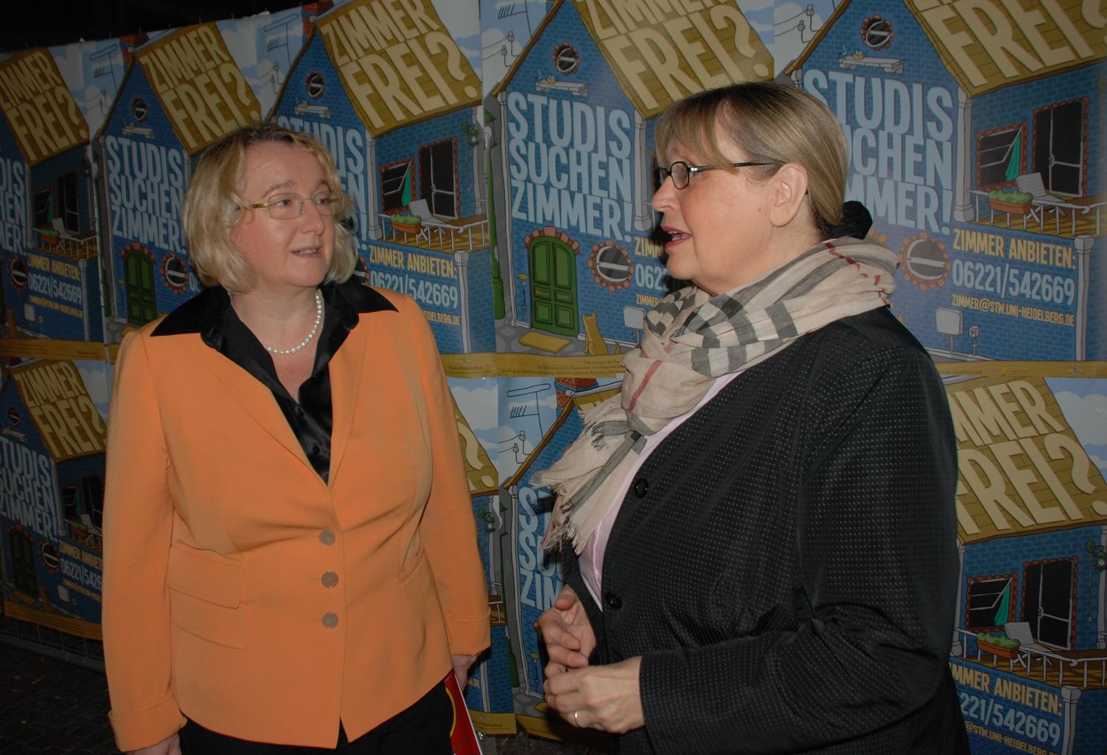 Theresia Bauer und Ulrike Leiblein im Gespräch vor einer Plakatwand zu "Studis suchen Zimmer"