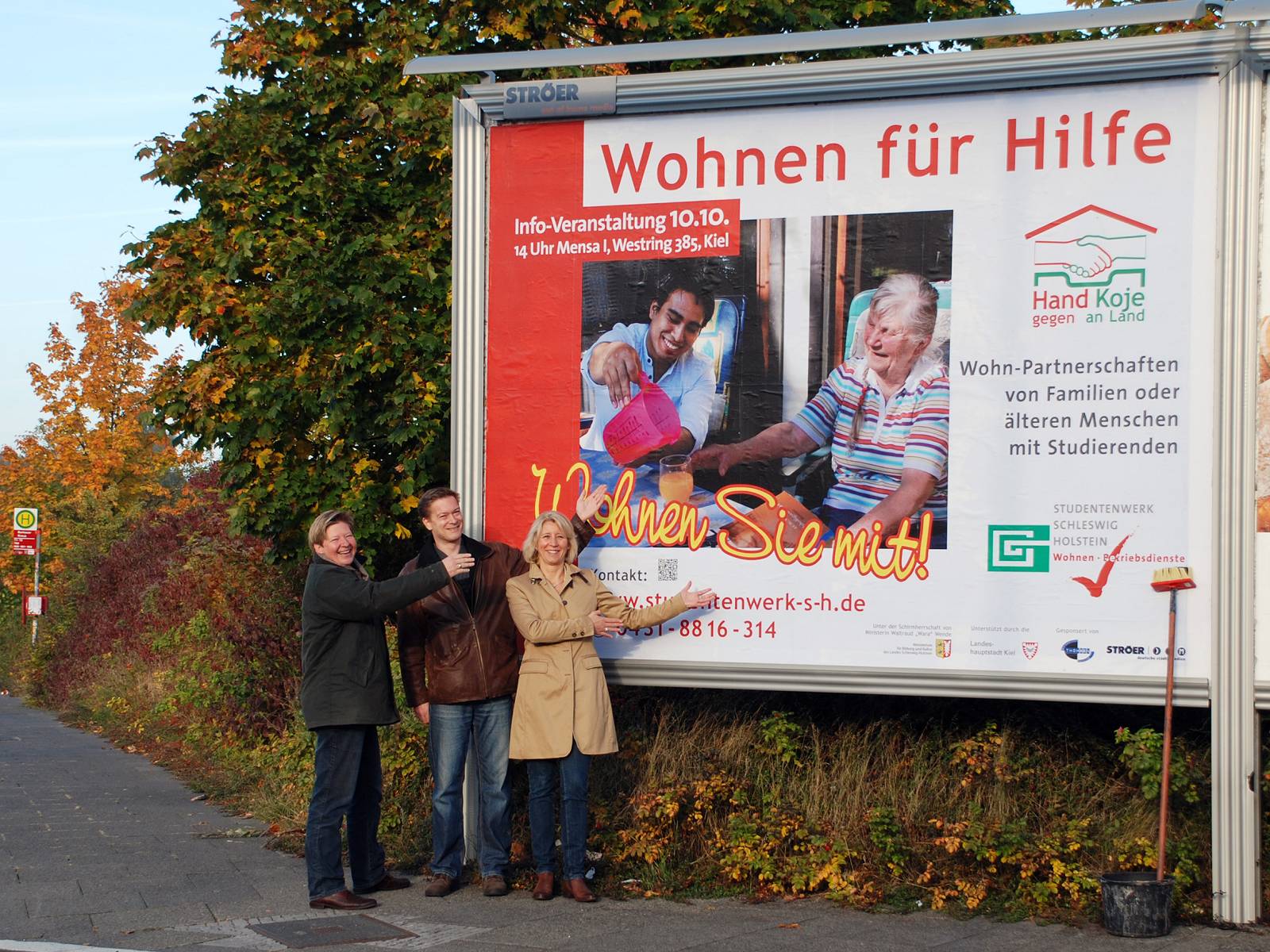 alt="Drei Personen stehen vor einer großen Plakatwerbewand und weisen stolz auf das Plakat mit Werbung für das Projekt Wohnen für Hilfe"