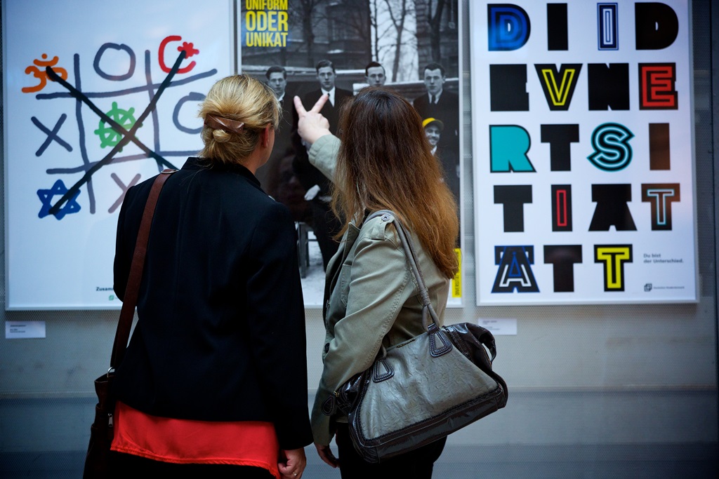 Zwei Mädchen stehen von drei Plakaten auf der Ausstellung und zeigen auf das Plakat in der Mitte.